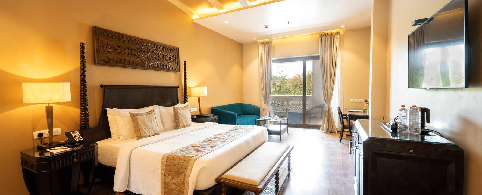 Premium Room at Essentia Luxury Resort & Spa, Udaipur showing a plush bed and an ottoman at its foot facing the TV over a table on the wall with a seating space by the door to the balcony