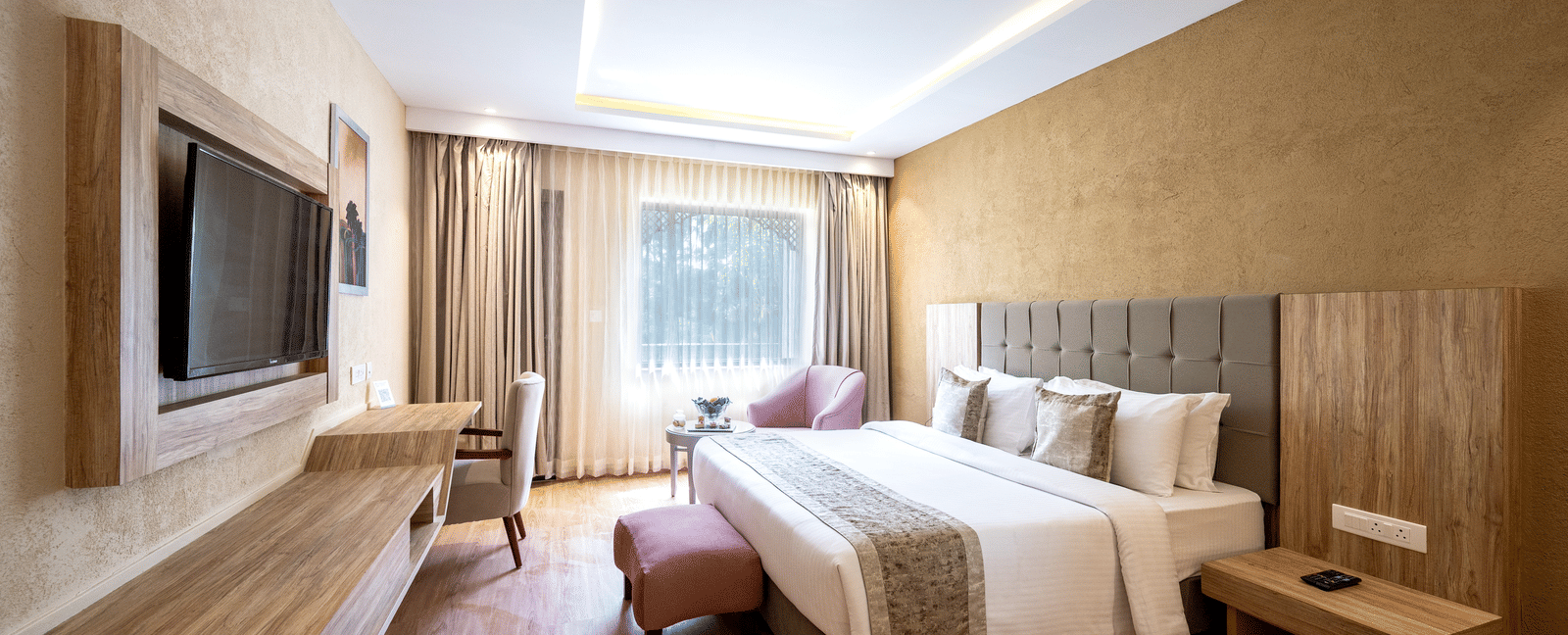 Standard Room at Essentia Luxury Resort & Spa, Udaipur featuring a plush bed witrh ottoman facing the TV on the wall over a running desk with a chair by the window with a seating area