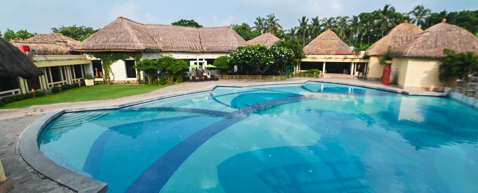 A daylight view of the common swimming pool at Vedic Village Spa Resort, featuring buildings with tatched roofs in the background.