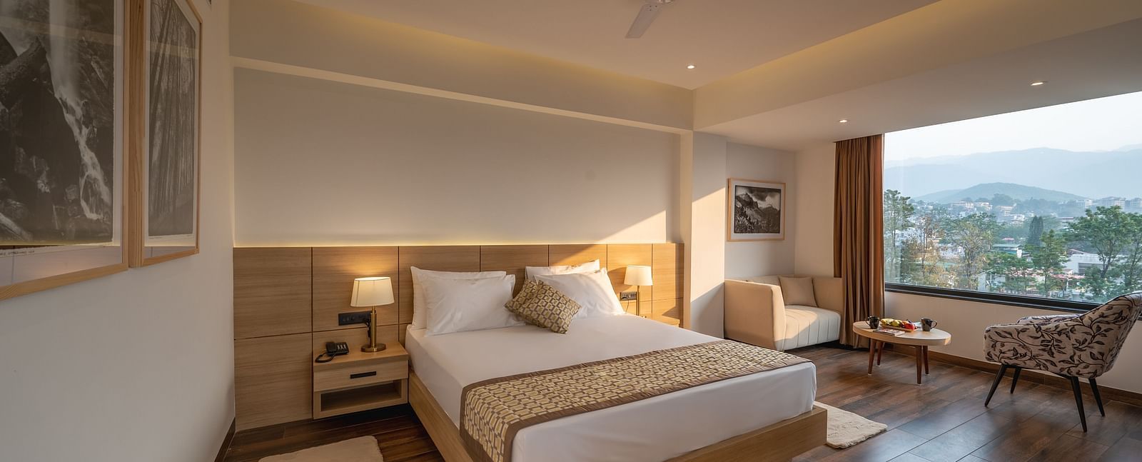 Full shot of Premium Room at Vishranti Hotel, Dehradun featuring a king bed with side tables, a TV, large window and bed lamps. 