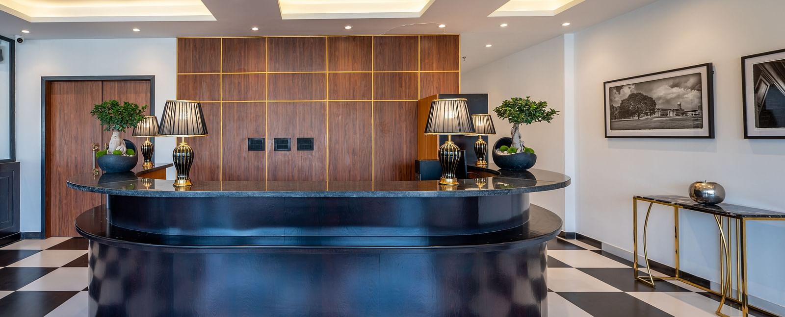  Reception area of Vishranti Hotel, Dehradun featuring a black reception desk with a curved design, a checkered black and white tiled floor, and a backdrop of wood-paneled walls.