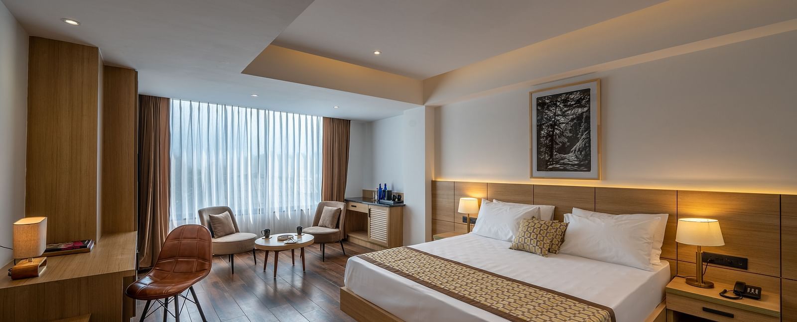 Shot of Premium Room at Vishranti Hotel, Dehradun featuring a king bed with side tables, a TV, large window and bed lamps. 