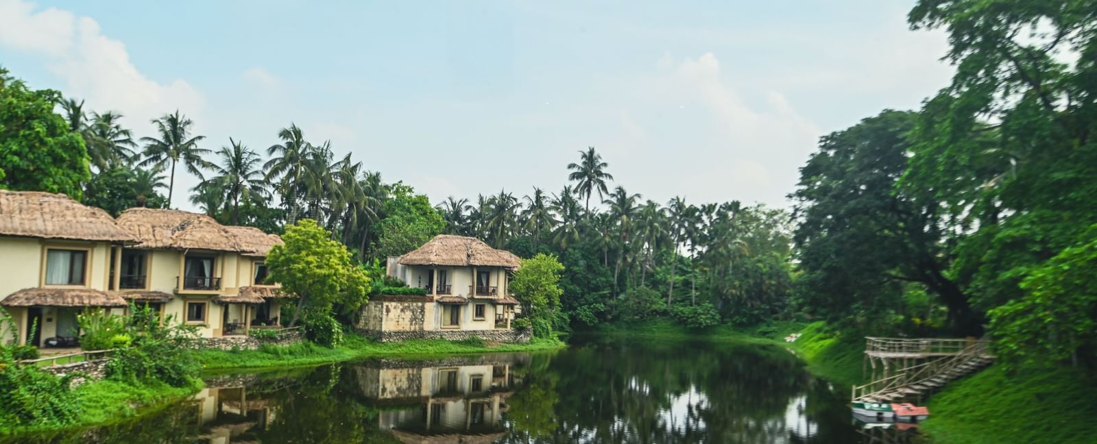 View of the lake at Vedic Village Spa Resort surrounded by greenery, with a boating deck on one side and building reflections visible under a blue sky.