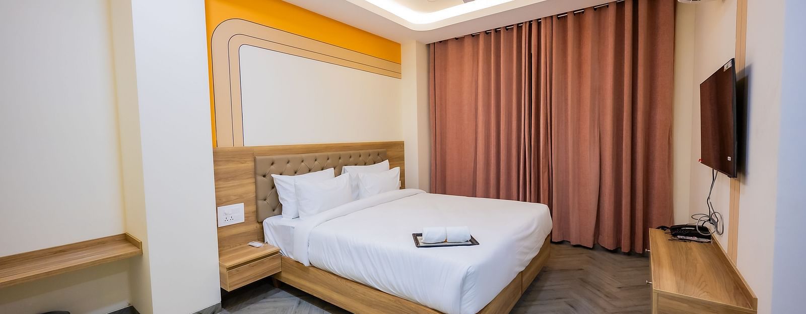 A cosy hotel room with soft lighting, a bed, curtains, and a TV mounted on the wall.