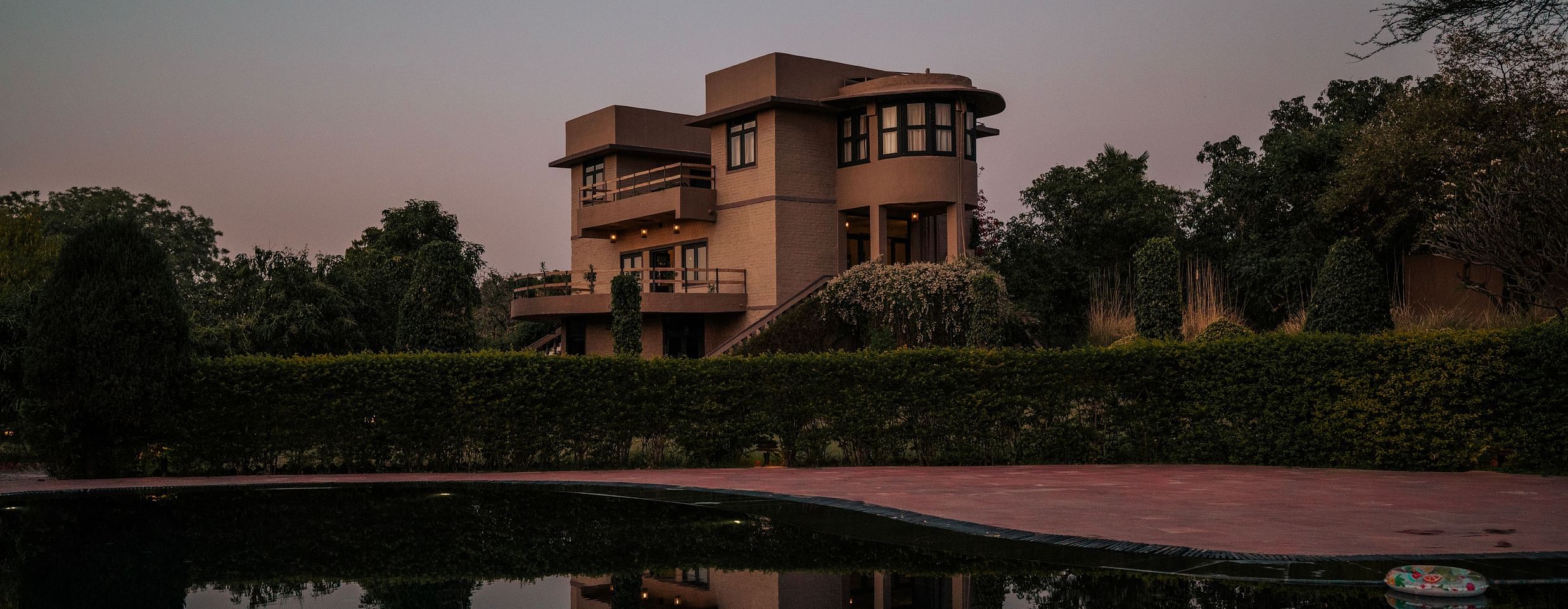 Stone building with multiple windows reflected in a still body of water. Trees and dusk sky in the background create a serene atmosphere - Clarks Safari, Pushkar