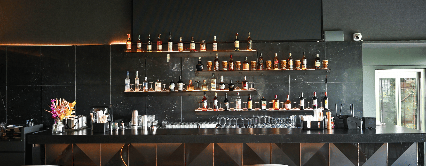 Modern bar at Hotel Daspalla, Hyderabad with black marble backdrop, premium spirit display on lit shelves, white bar stools, and curved wooden ceiling beams with warm lighting.