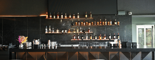 Modern bar at Hotel Daspalla, Hyderabad with black marble backdrop, premium spirit display on lit shelves, white bar stools, and curved wooden ceiling beams with warm lighting.