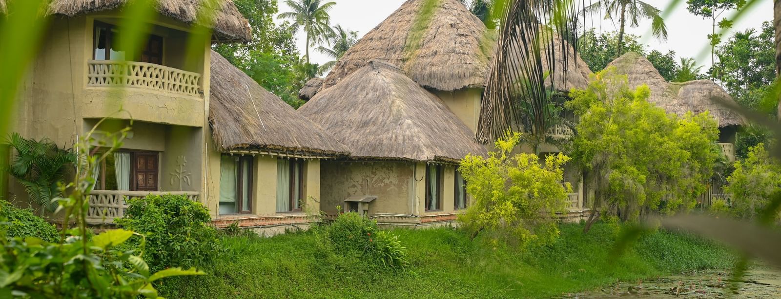 Thatched-roof Earth Villas at Vedic Village Spa Resort overlooking a water body, with lush green vegetation lining the perimeter of the natural surroundings.