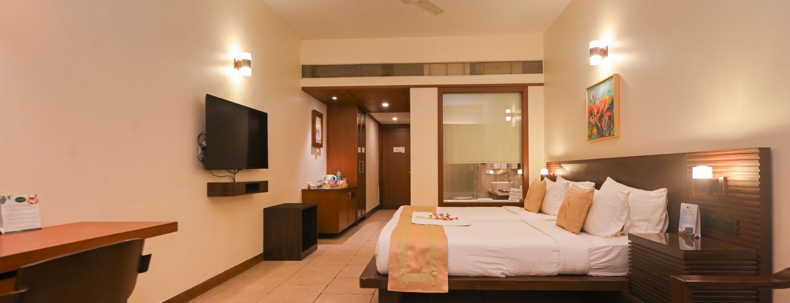 Spacious view of the Standard Suite at Vedic Village Spa Resort with a double bed, flat-screen TV, work desk with chair, glass-topped table, and wooden furnishings.
