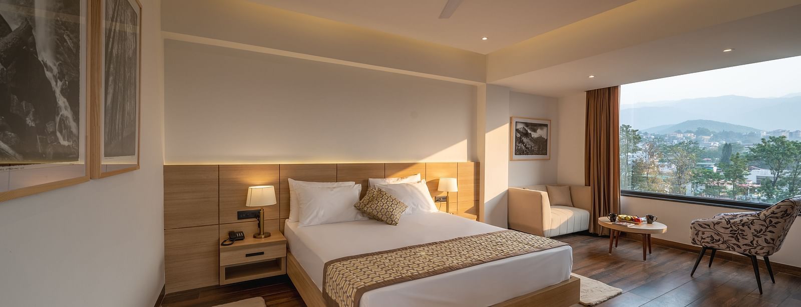 Full shot of Premium Room at Vishranti Hotel, Dehradun featuring a king bed with side tables, a TV, large window and bed lamps. 