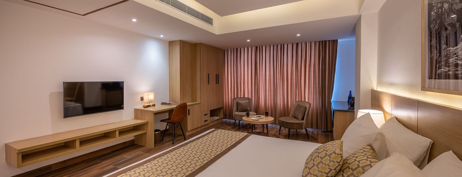 Side Shot of Premium Room at Vishranti Hotel, Dehradun featuring a king bed with side tables, a TV, large window and bed lamps. 