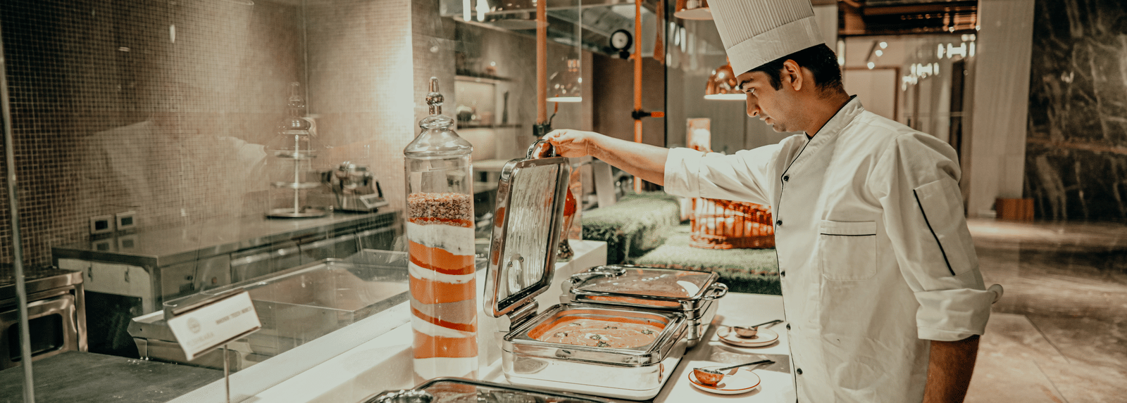 A chef in a white uniform prepares food behind a counter in a professional kitchen. There are different dishes in metal containers on the counter.