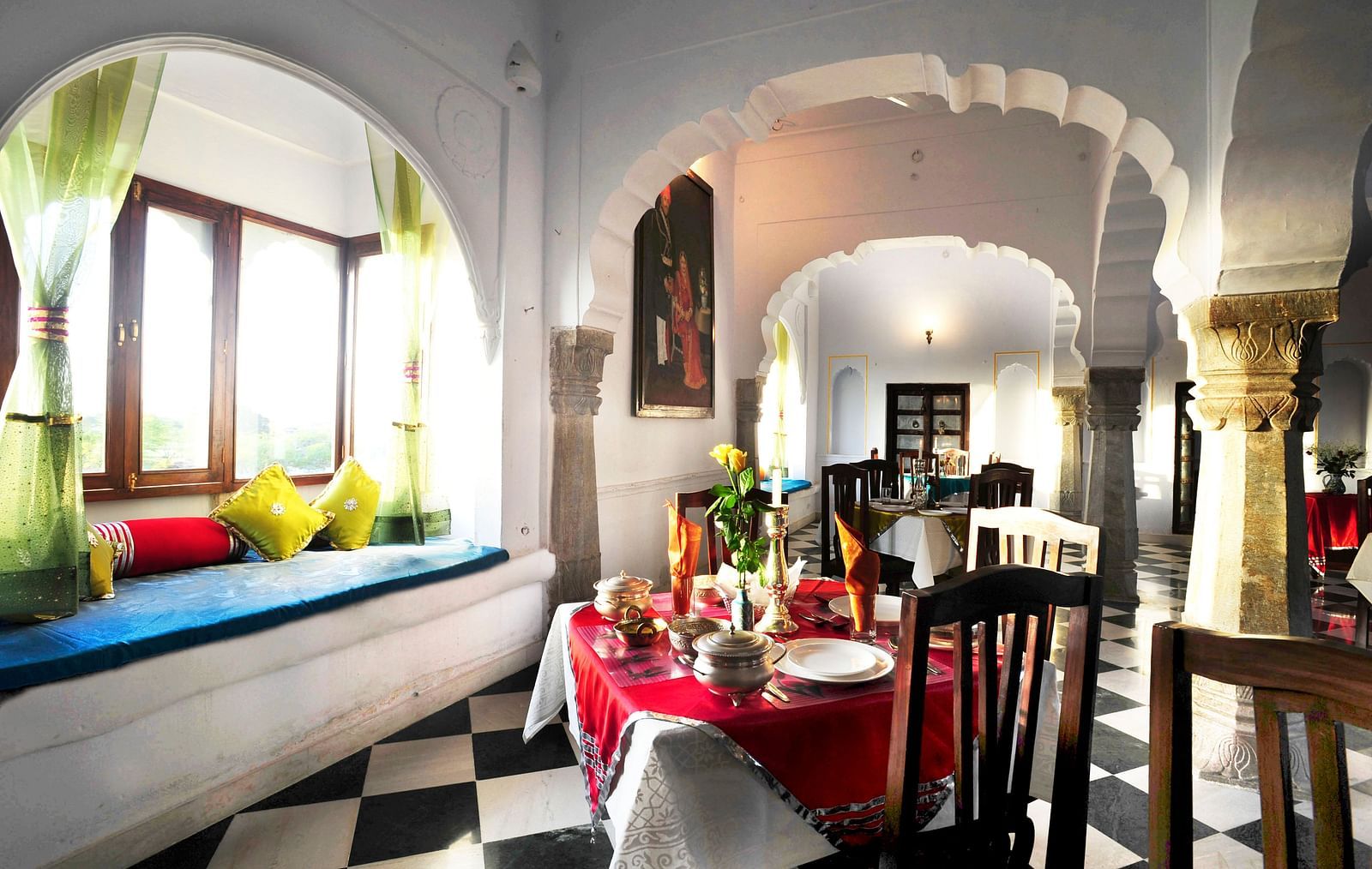A bright dining area at Fort Barli, featuring arched doorways, checkered floors, and a window seat, blends architectural charm with a cosy meal setting.