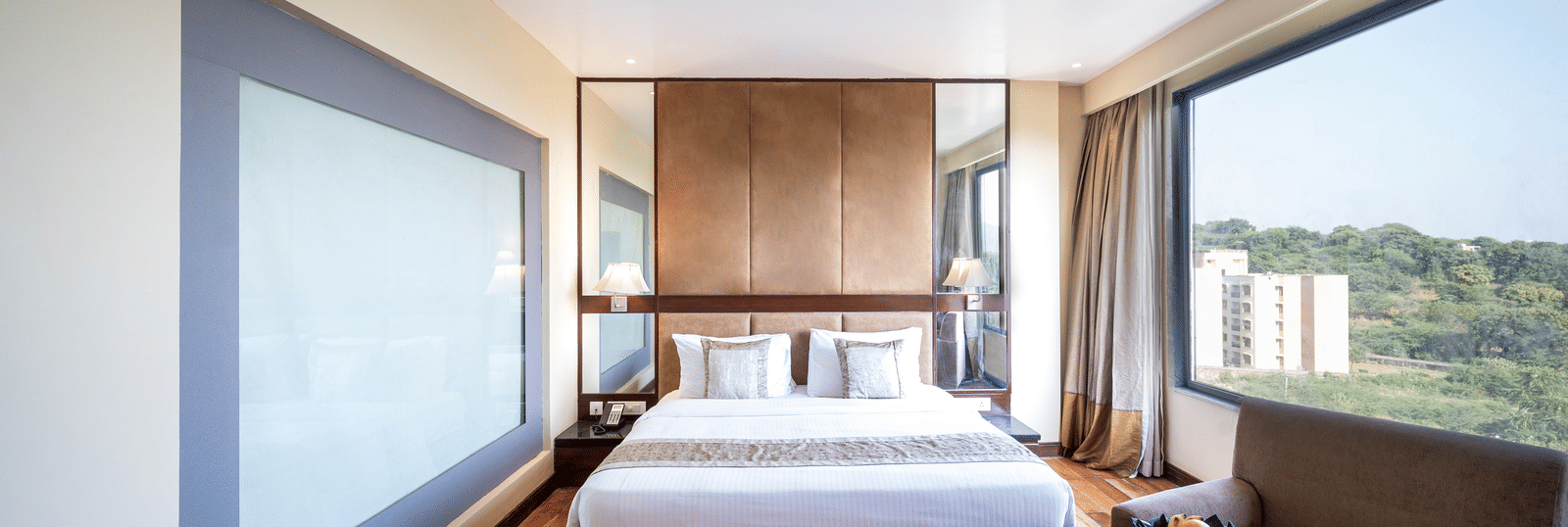 Premium Room at Essentia Luxury Resort & Spa, Udaipur featuring a plush bed by a larg window with the view of trees and a seating space near the window