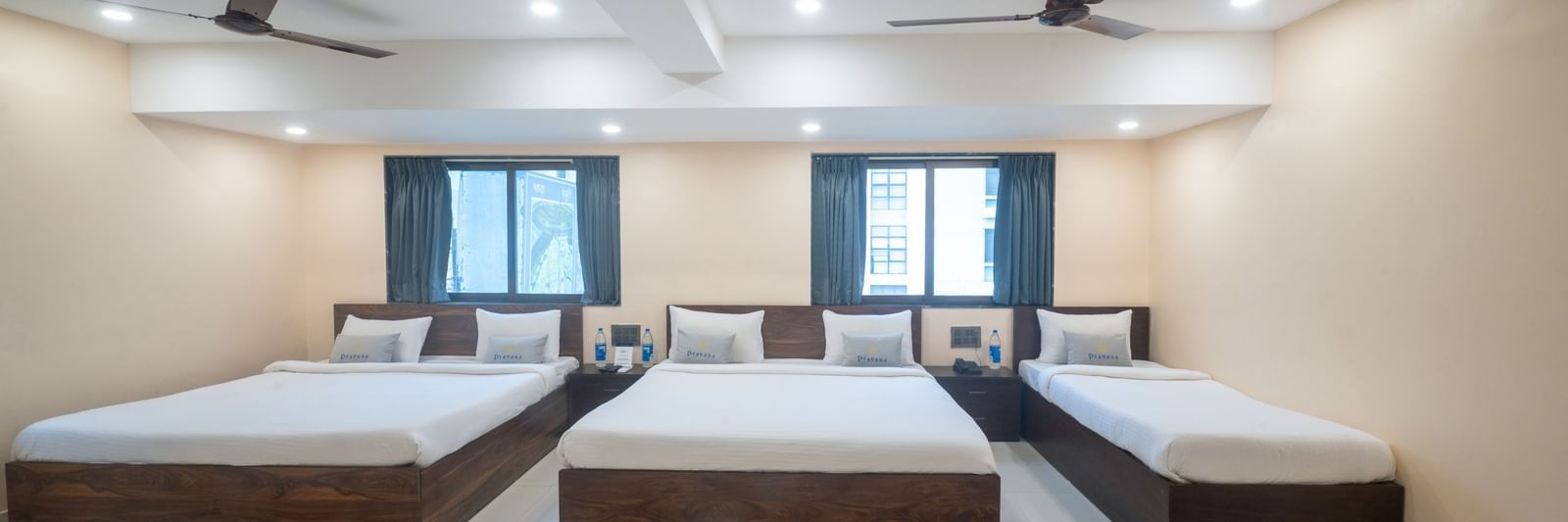 A large room at Pravasa Stay, Shirdi, contains three beds, two ceiling fans, and multiple windows.
