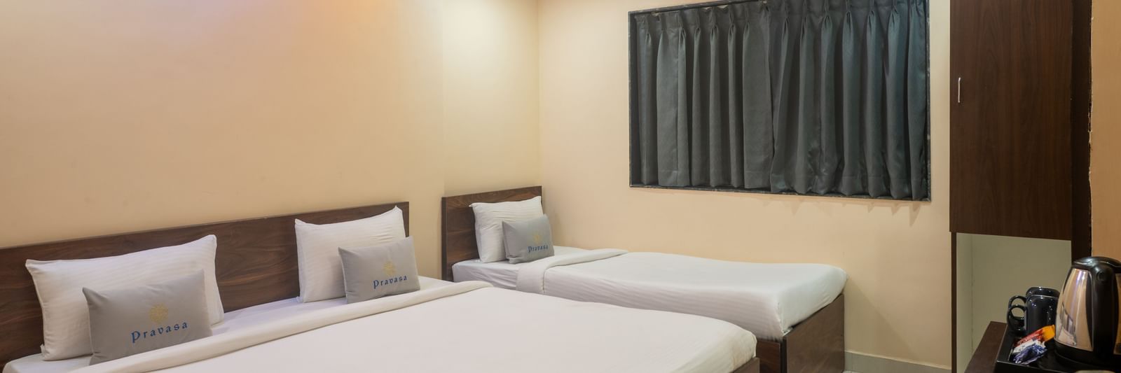 A room at Pravasa Stay, Shirdi, is equipped with one double bed and one single bed with a dark wardrobe nearby.