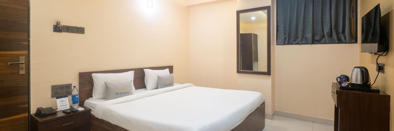 A room at Pravasa Stay, Shirdi, is furnished with a wooden bed, a bedside table, and a wall-mounted mirror.