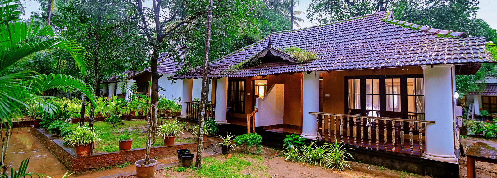 A tropical resort cottage with a dark tiled roof and small wooden balcony is surrounded by thick foliage, trees, and a patchy lawn.