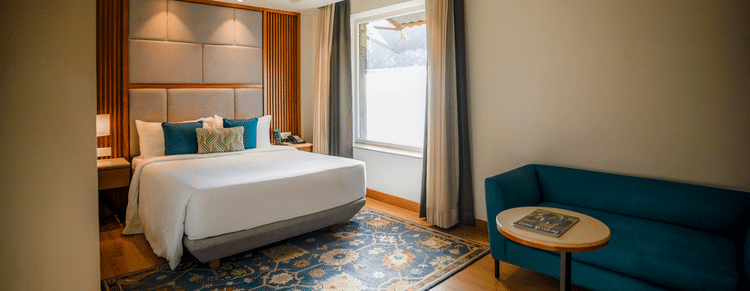 A brightly lit hotel room features a neatly made bed with white linens, a teal armchair, and a small round side table with a lamp. A window with sheer curtains allows natural light to filter in - Ananta Spa and Resort, Pushkar