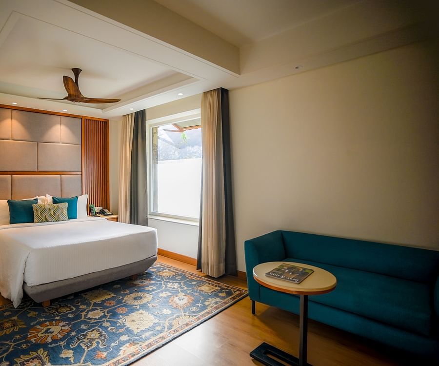 A brightly lit hotel room features a neatly made bed with white linens, a teal armchair, and a small round side table with a lamp. A window with sheer curtains allows natural light to filter in - Ananta Spa and Resort, Pushkar