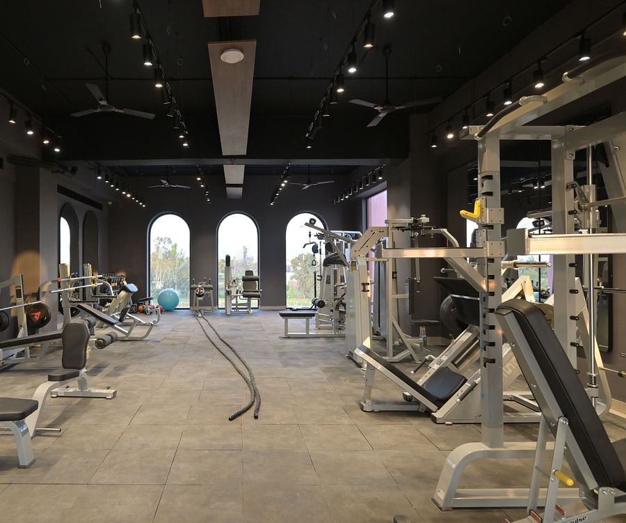 The interior of a well-equipped gym with various weightlifting and exercise machines, set against a backdrop of arched windows and a dark ceiling with recessed lighting. - Clarks Safari Haridwar