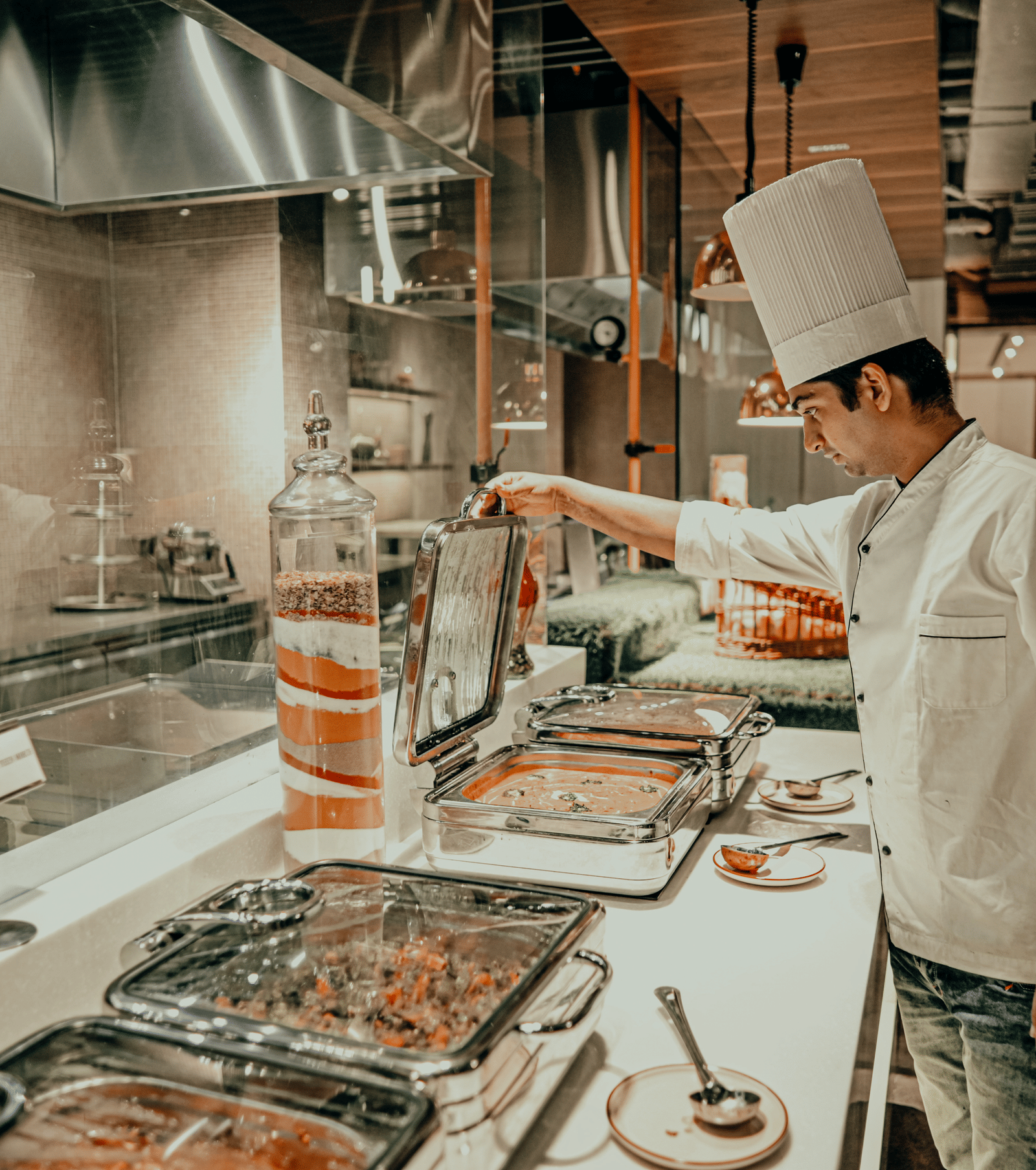A chef in a white uniform prepares food behind a counter in a professional kitchen. There are different dishes in metal containers on the counter.