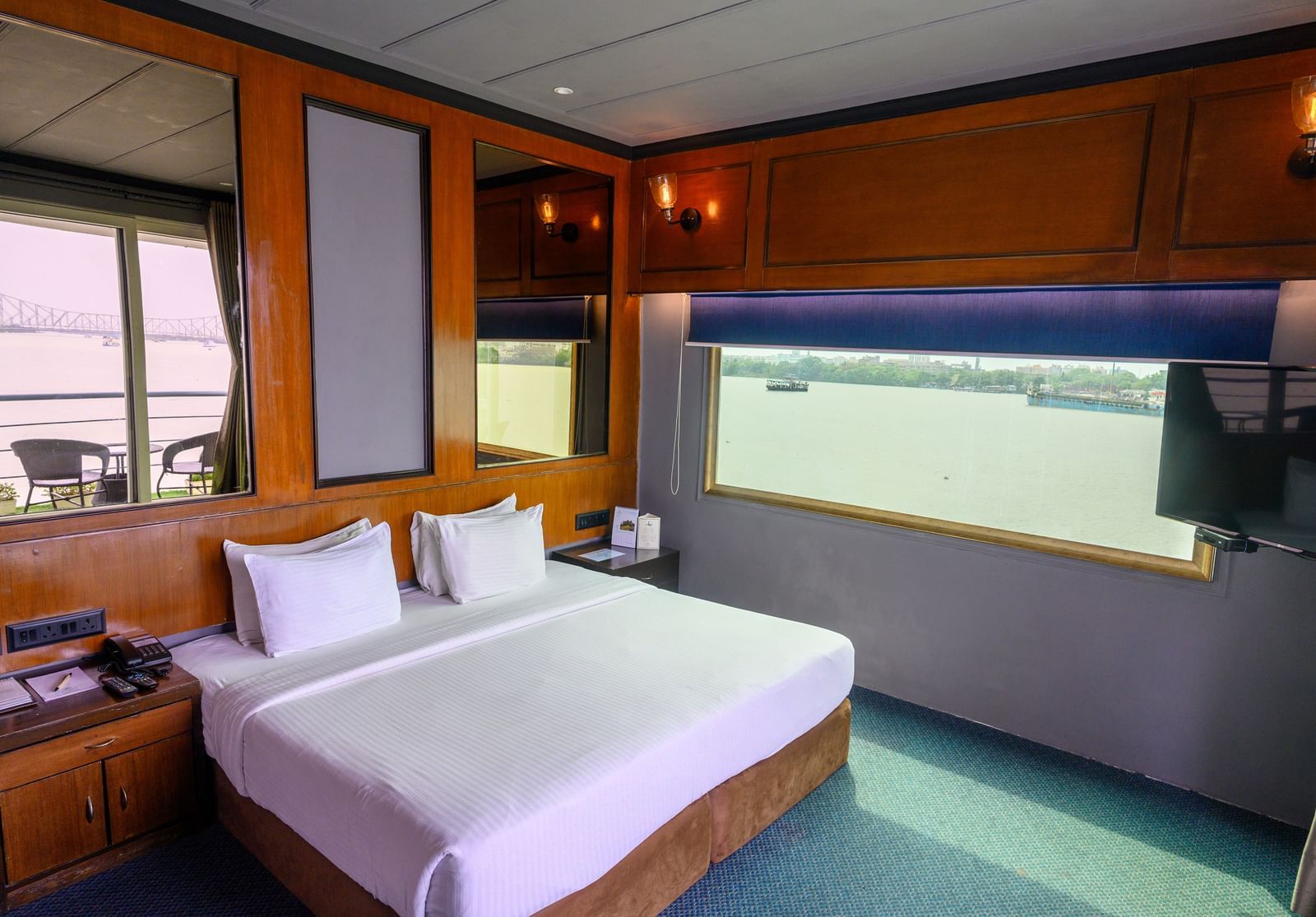 A spacious hotel room at Polo Floatel Kolkata with a large bed in the center. The room has a window with a unique, artistic frame that offers a view of the river and a nearby dock.