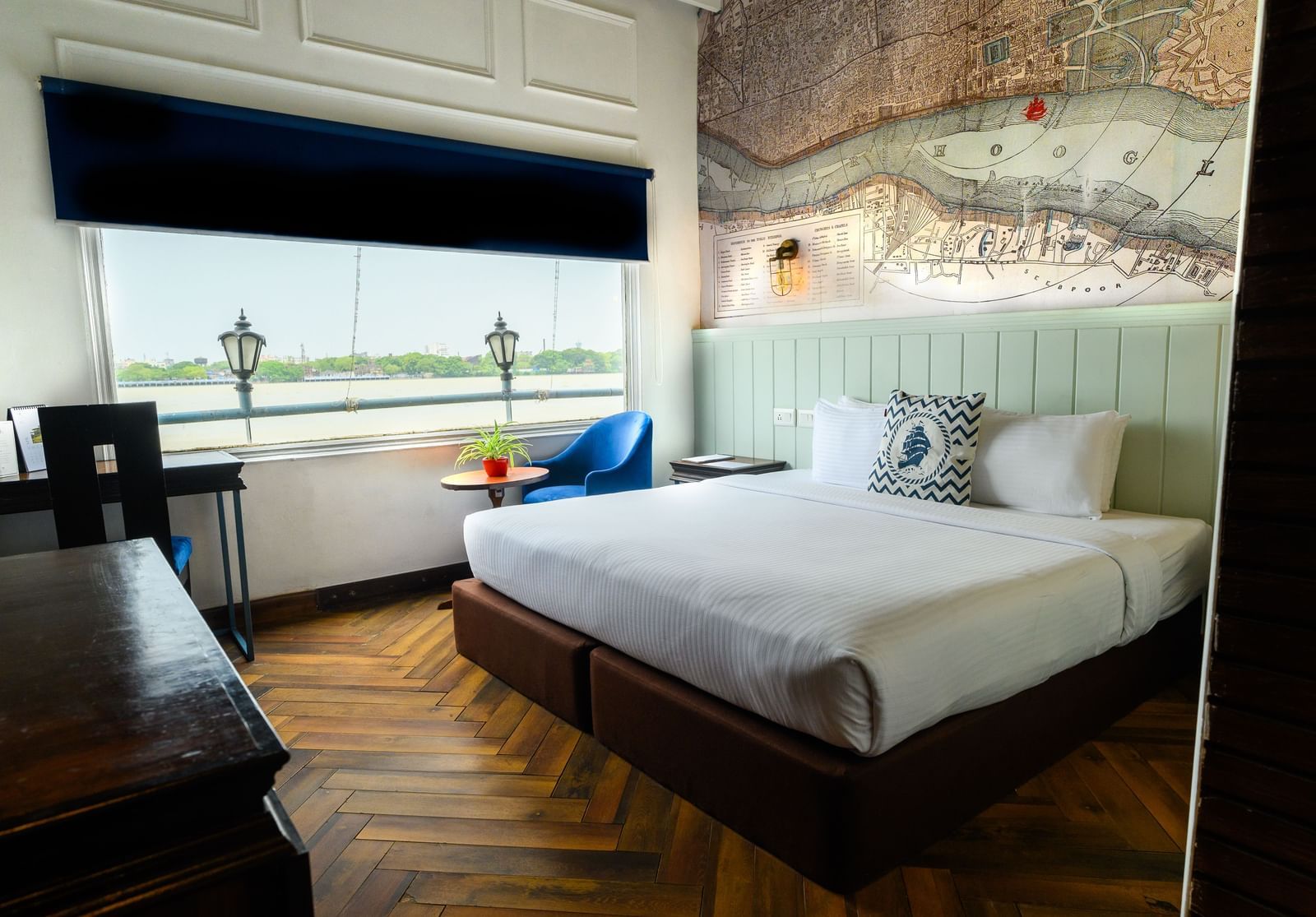 An elegant hotel room at Polo Floatel Kolkata featuring a large bed with a white duvet. The room has wooden paneling and a long window that provides a panoramic view of the river, with a small seating area and a television.