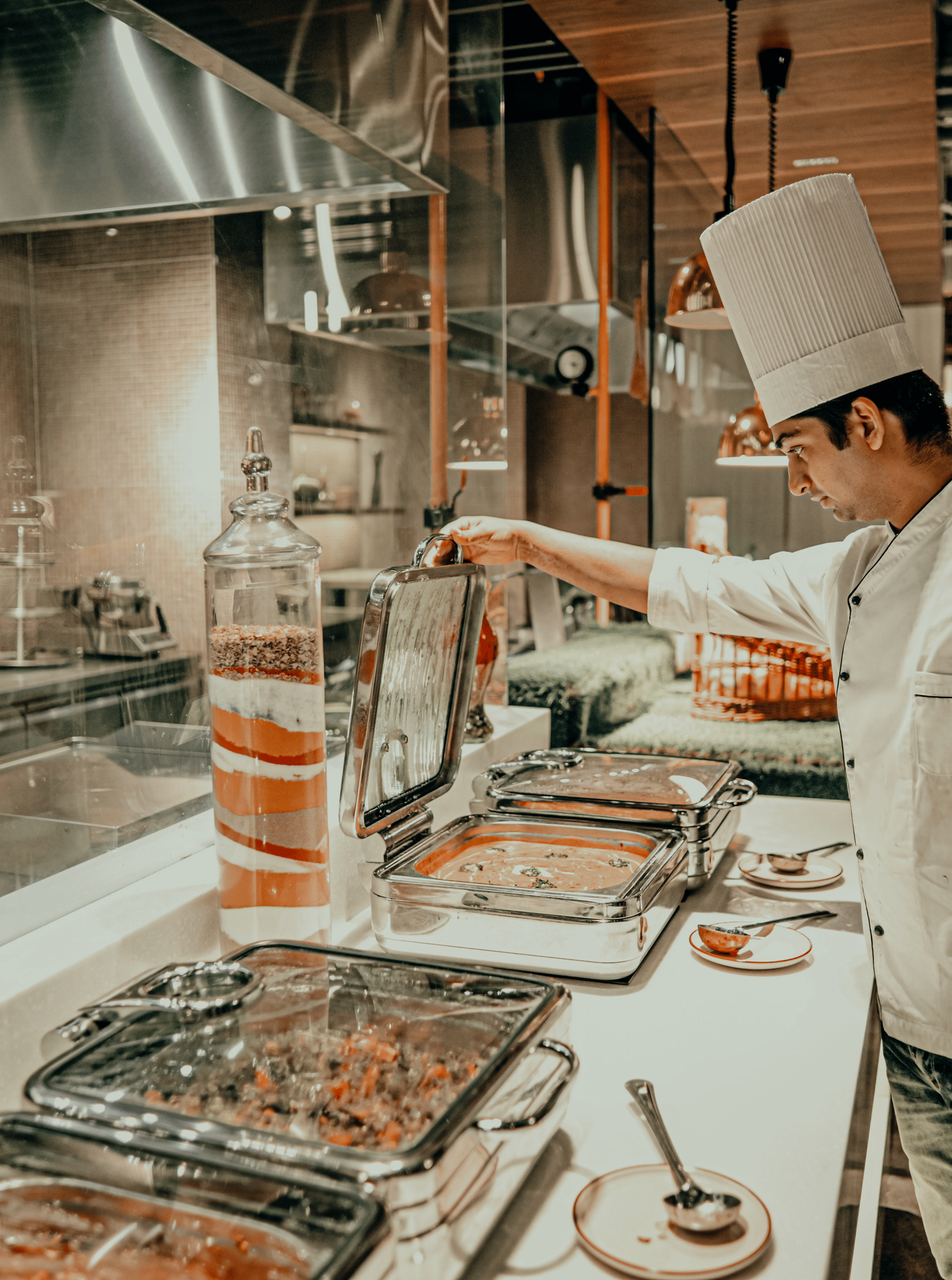 A chef in a white uniform prepares food behind a counter in a professional kitchen. There are different dishes in metal containers on the counter.