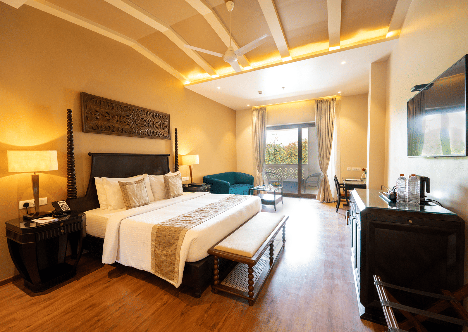 Premium Room at Essentia Luxury Resort & Spa, Udaipur showing a plush bed and an ottoman at its foot facing the TV over a table on the wall with a seating space by the door to the balcony