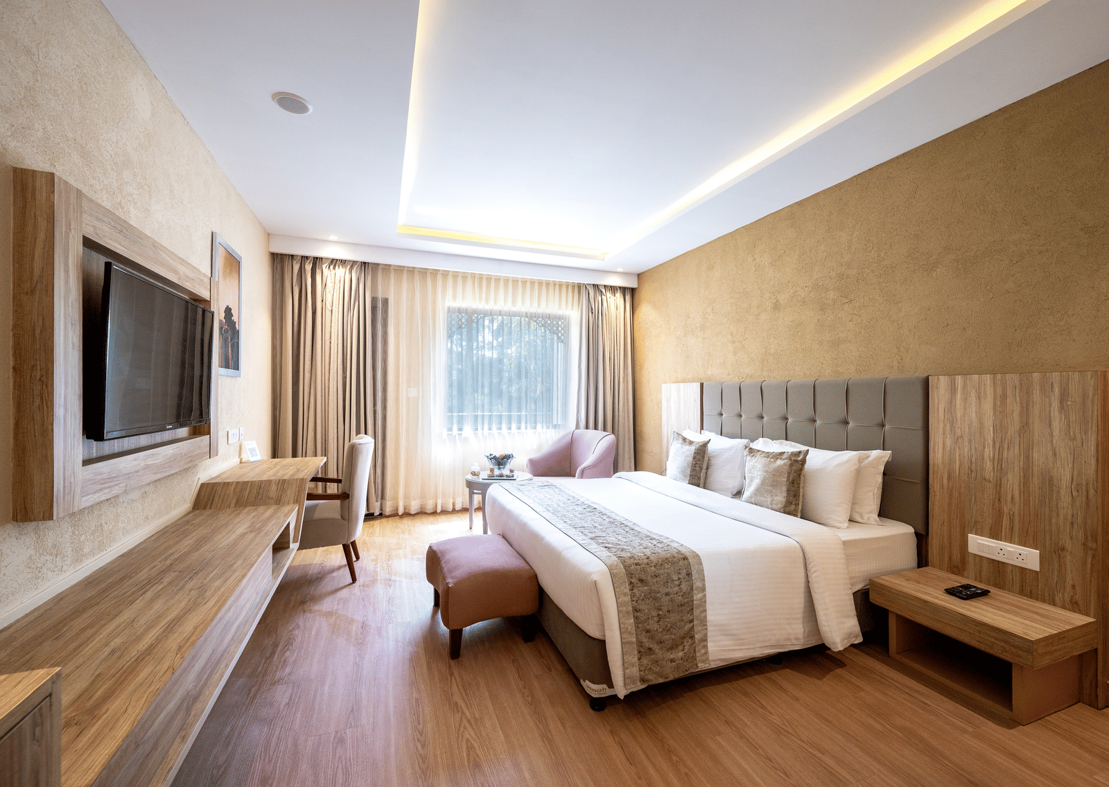 Standard Room at Essentia Luxury Resort & Spa, Udaipur featuring a plush bed witrh ottoman facing the TV on the wall over a running desk with a chair by the window with a seating area