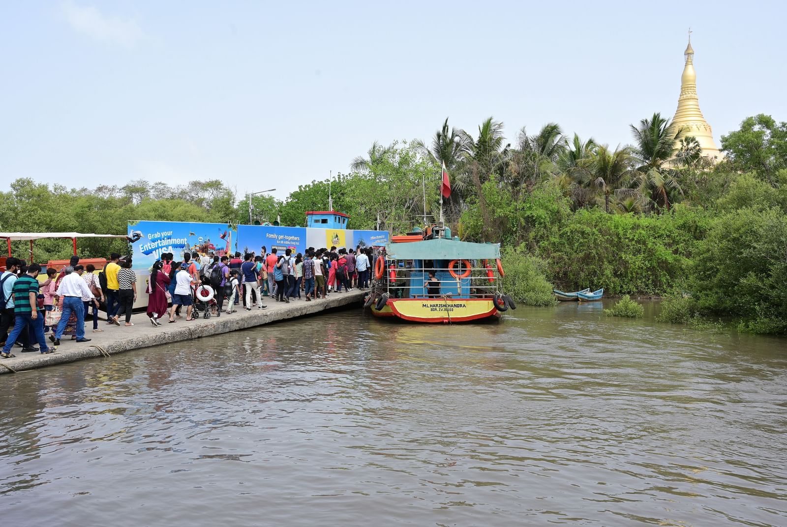 Boat docked on a river with a crowd of people boarding, against a blue sky background.