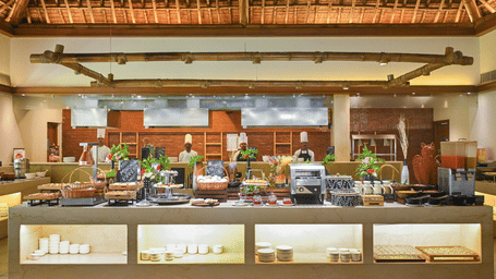 Restaurant buffet spread at Vedic Village Spa Resort Kolkata with a long counter displaying multiple food dishes under a high exposed wooden thatched roof ceiling.