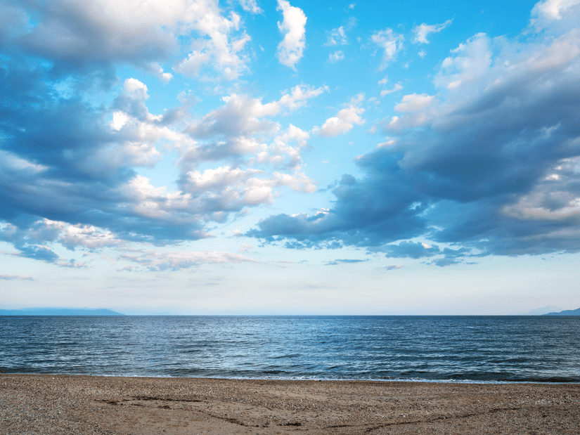 Wide open seascape view with a sandy shoreline in the foreground calm blue sea in the middle and a dramatic sky filled with white and grey clouds stretching across the horizon.