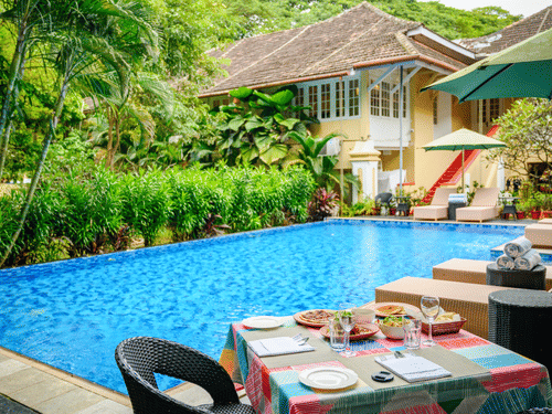 A dining table set by the swimming pool at The Tower House, surrounded by greenery, overlooking the buildings in the property