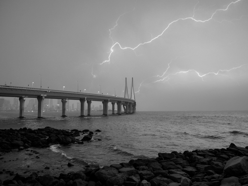 Black and white image of a long bridge extending over the sea with lightning striking in the cloudy sky above and rocky shoreline in the foreground.