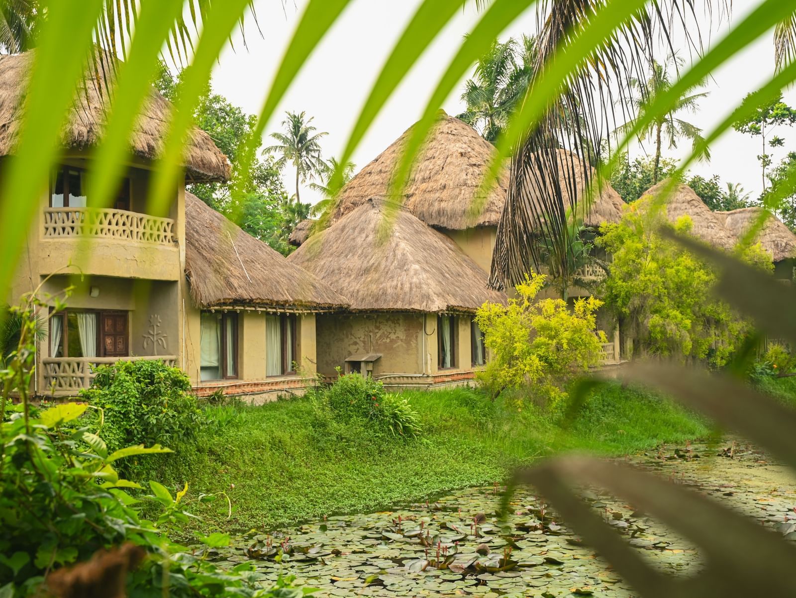 Exterior view of Earth Villa at Vedic Village Spa Resort showing mud-built structures with thatched roofs, set beside a calm water body and surrounded by dense greenery.