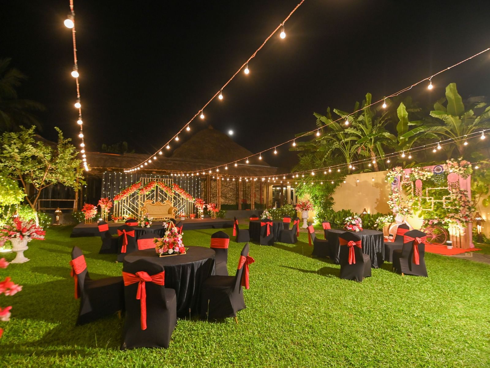 A night time view of a neatly trimmed lawn space at Vedic Village Spa Resort, featuring warm decorative lightsand black banquet chairs with red bows.