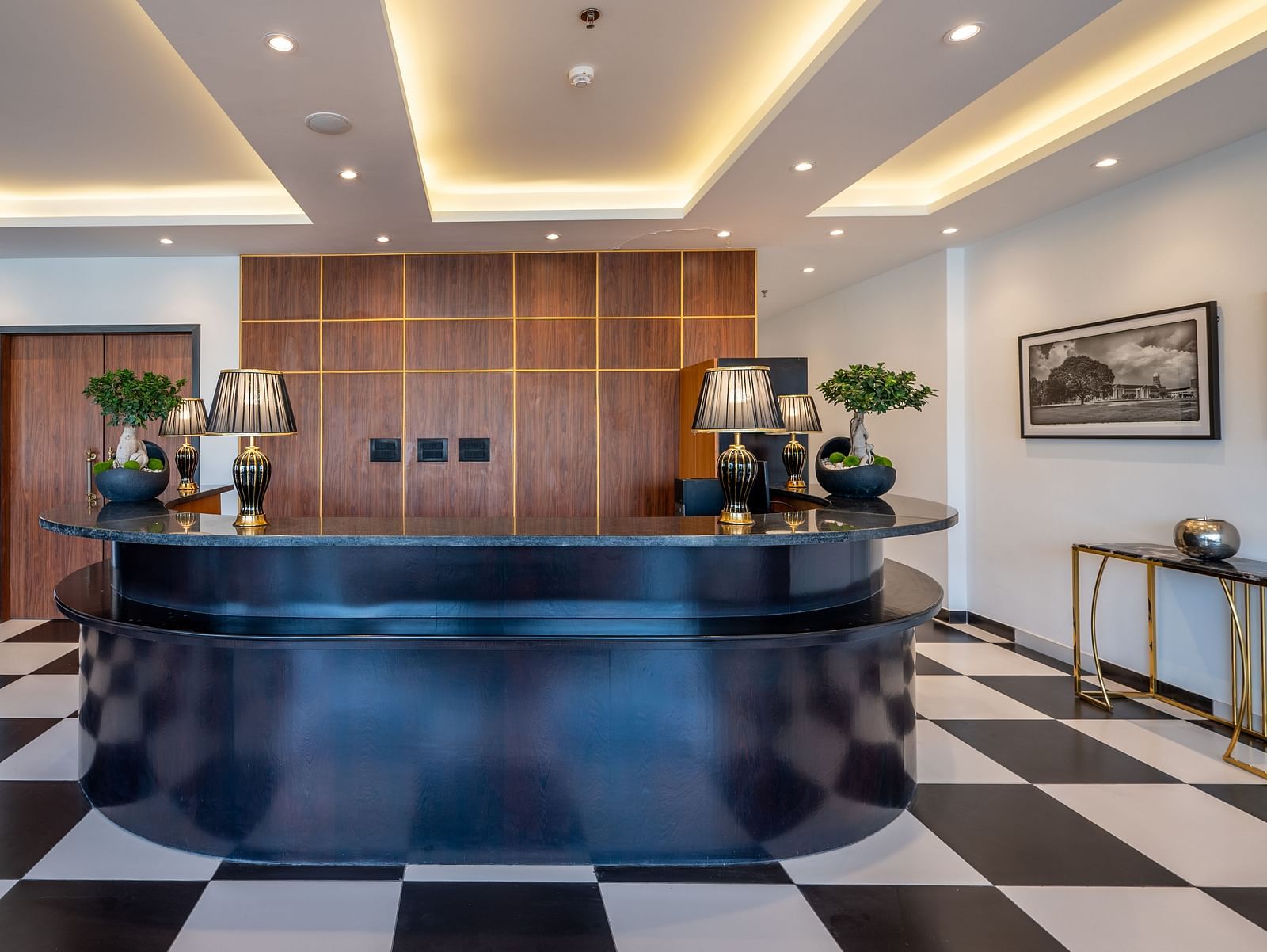  Reception area of Vishranti Resort, Dehradun featuring a black reception desk with a curved design, a checkered black and white tiled floor, and a backdrop of wood-paneled walls.