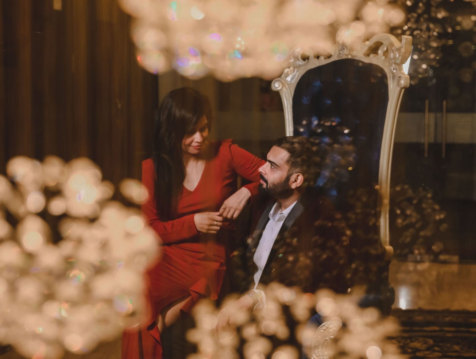 A couple, the woman in a red dress and the man in a suit, are seated intimately together in a dimly lit room with decorative lighting elements in the foreground and background.