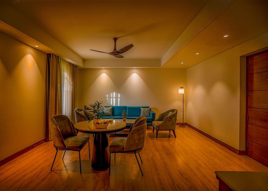 A cozy hotel suite includes a small round dining table with three chairs and a separate seating area with a teal sofa and a coffee table. Warm lighting from wall sconces illuminates the room - Ananta Spa and Resort, Pushkar