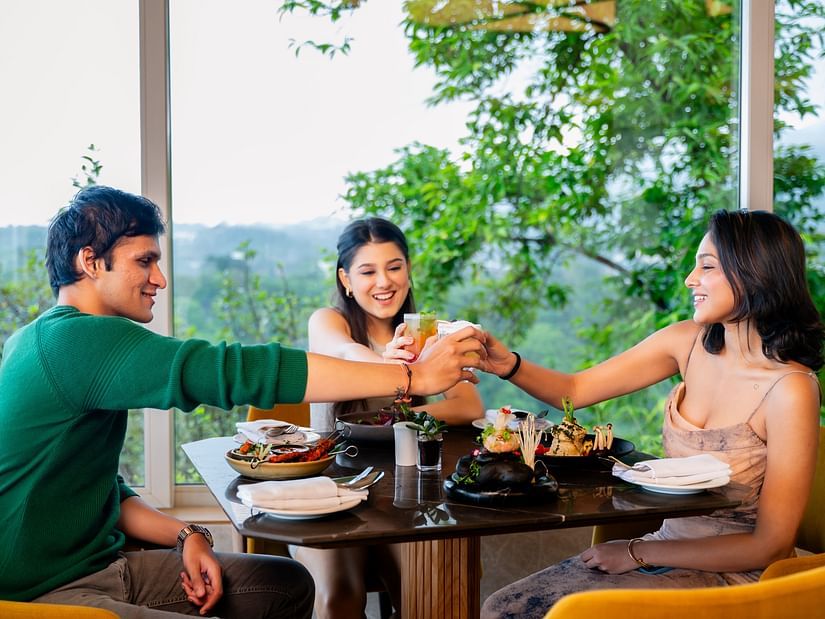 An image of three people sitting in a cafe clinging there glasses together with an amazing view behind them - The Dukes Retreat, Khandala