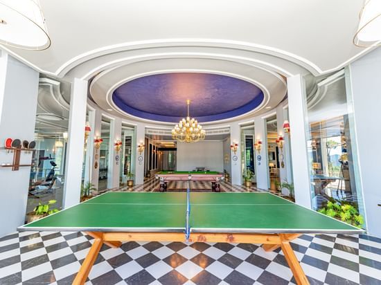 Grand indoor recreational room at The Manor Luxury Apartments, Shimla, featuring a table tennis, checkered tile floor, elegant chandelier, and mirrored walls, creating a luxurious leisure space.