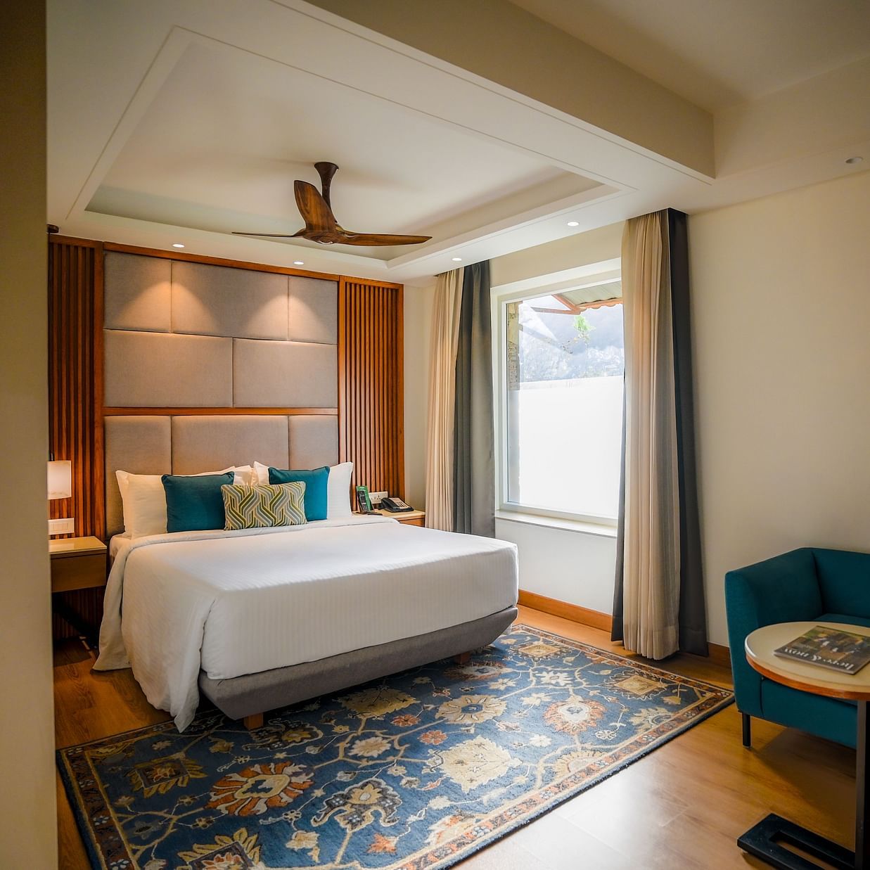 A brightly lit hotel room features a neatly made bed with white linens, a teal armchair, and a small round side table with a lamp. A window with sheer curtains allows natural light to filter in - Ananta Spa and Resort, Pushkar