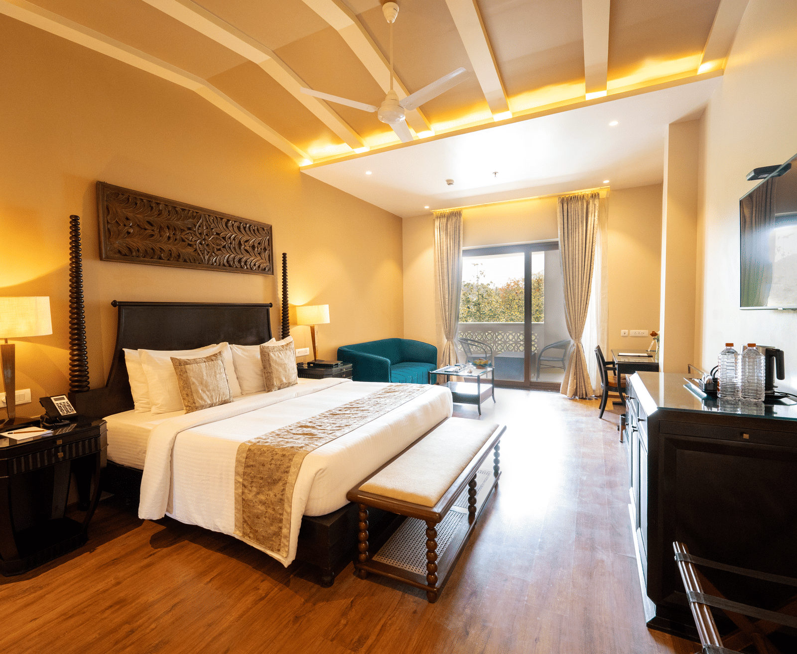Premium Room at Essentia Luxury Resort & Spa, Udaipur showing a plush bed and an ottoman at its foot facing the TV over a table on the wall with a seating space by the door to the balcony