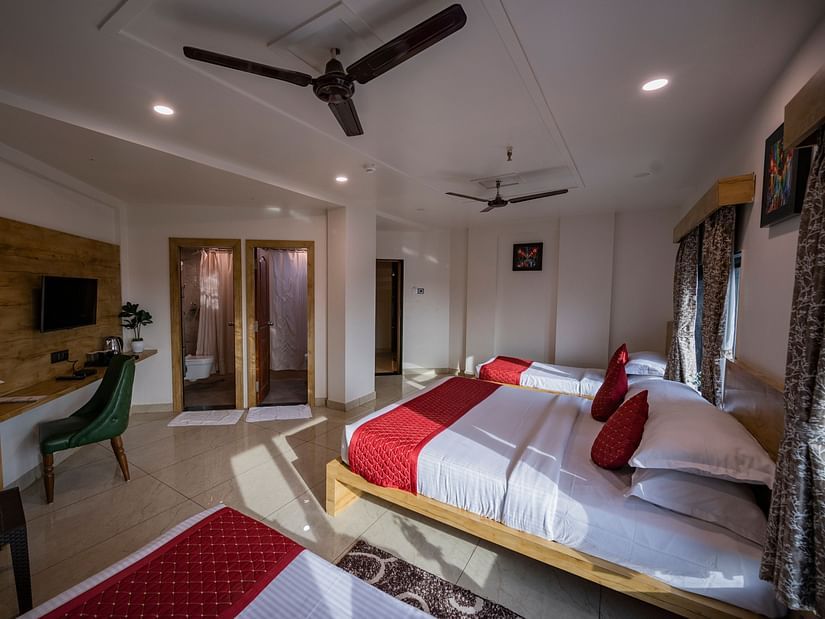 A room at Clarks Collection Somnath with beds, red runners, desk, tv, ceiling fan, chair, table and fruits on the table and wooden decor.