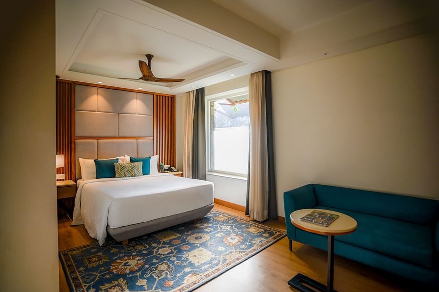 A brightly lit hotel room features a neatly made bed with white linens, a teal armchair, and a small round side table with a lamp. A window with sheer curtains allows natural light to filter in - Ananta Spa and Resort, Pushkar