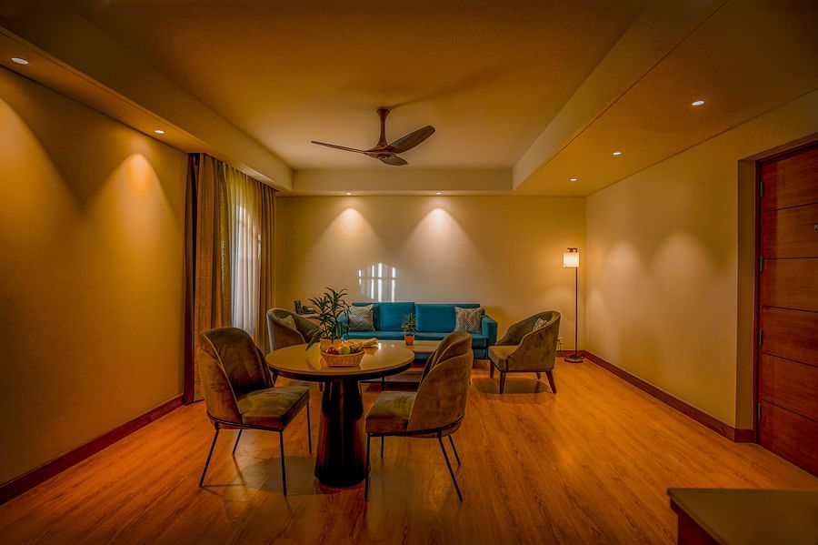 A cozy hotel suite includes a small round dining table with three chairs and a separate seating area with a teal sofa and a coffee table. Warm lighting from wall sconces illuminates the room - Ananta Spa and Resort, Pushkar