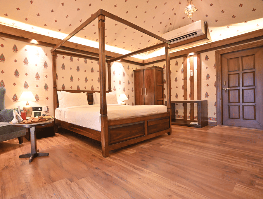 A Premium Tent featuring a poster bed, air conditioner, wardrobe, bedside table with lamp, and a sofa chair with centre table where food items are kept - The Baagh Ananta Elite, Ranthambore