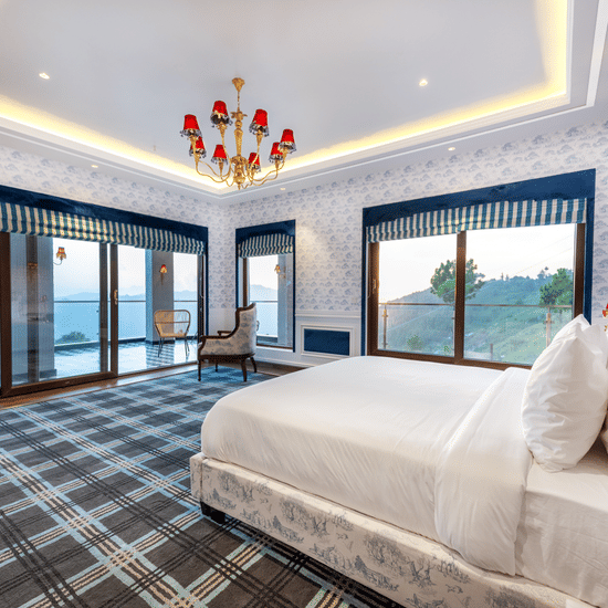 A bright, elegant interior of the Presidential Suite at The Manor Sports & Wellness Hotel featuring a large white bed, plaid carpet, statement chandelier, and floor-to-ceiling windows with mountain and lake views.